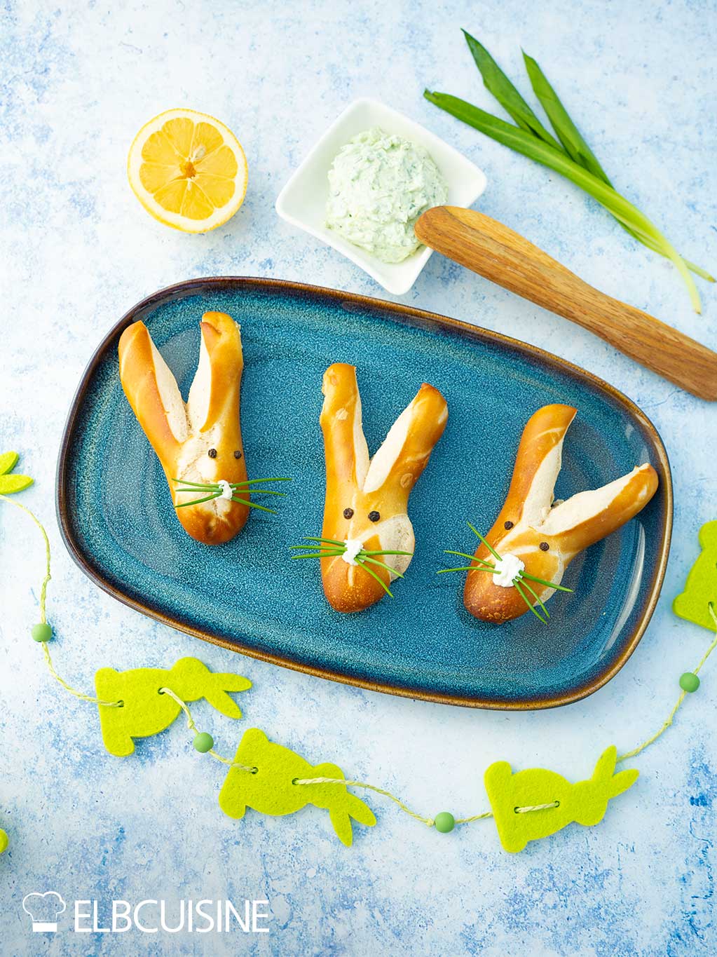 Ein Platte mit goldbraun gebackener Laugenbrötchen in niedlicher Hasenform mit langen Ohren und einer Nase aus Frischkäse und Schnurrhaare aus Schnittlauch mit einem hellgrünen Bärlauch-Dip bereit für den Osterbrunch.