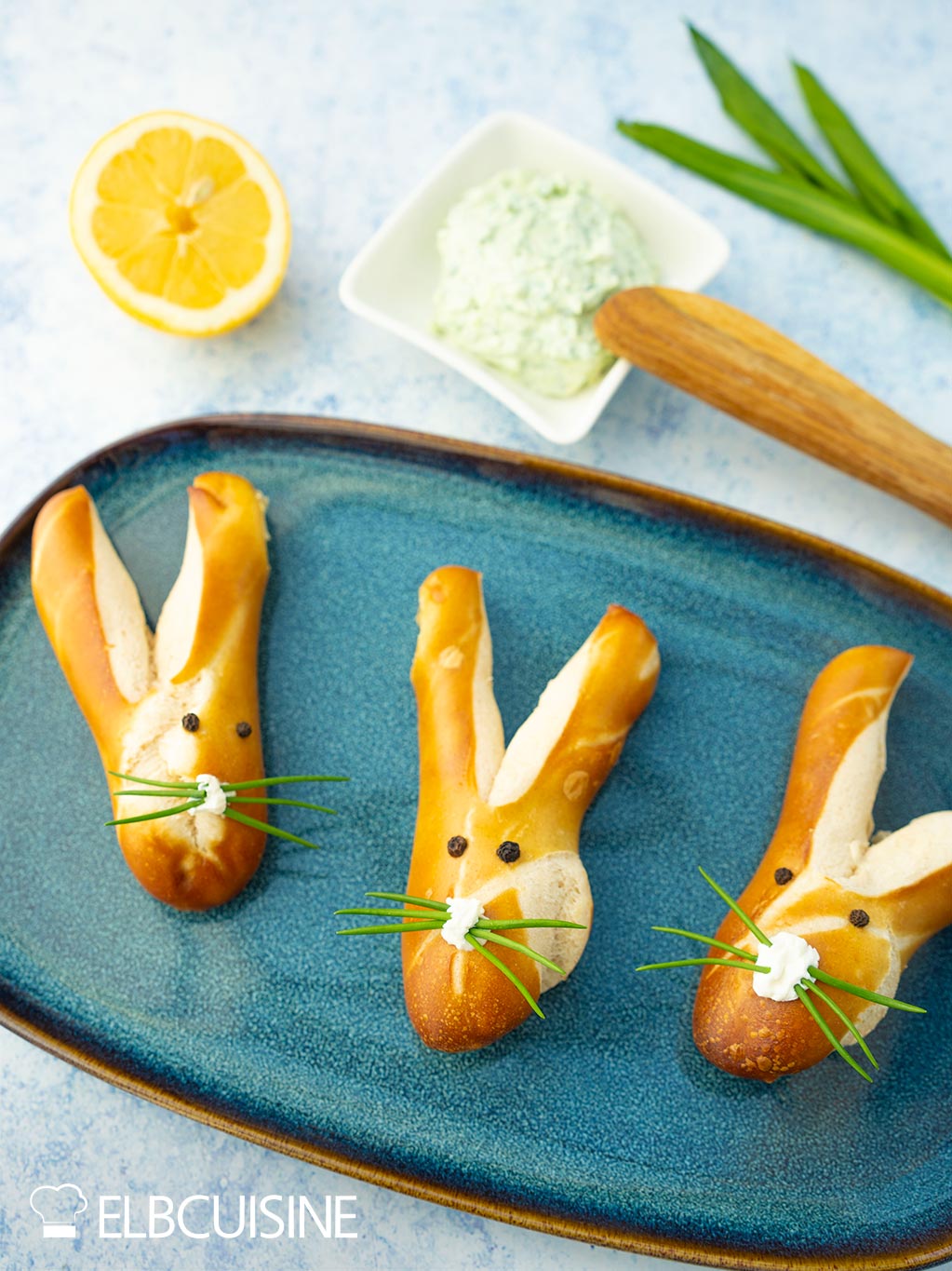 Ein Platte mit goldbraun gebackener Laugenbrötchen in niedlicher Hasenform mit langen Ohren und einer Nase aus Frischkäse und Schnurrhaare aus Schnittlauch mit einem hellgrünen Bärlauch-Dip bereit für den Osterbrunch.
