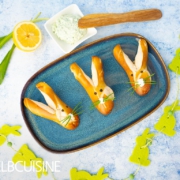 Ein Platte mit goldbraun gebackener Laugenbrötchen in niedlicher Hasenform mit langen Ohren und einer Nase aus Frischkäse und Schnurrhaare aus Schnittlauch mit einem hellgrünen Bärlauch-Dip bereit für den Osterbrunch.
