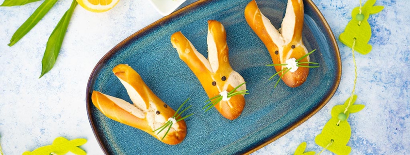 Ein Platte mit goldbraun gebackener Laugenbrötchen in niedlicher Hasenform mit langen Ohren und einer Nase aus Frischkäse und Schnurrhaare aus Schnittlauch mit einem hellgrünen Bärlauch-Dip bereit für den Osterbrunch.