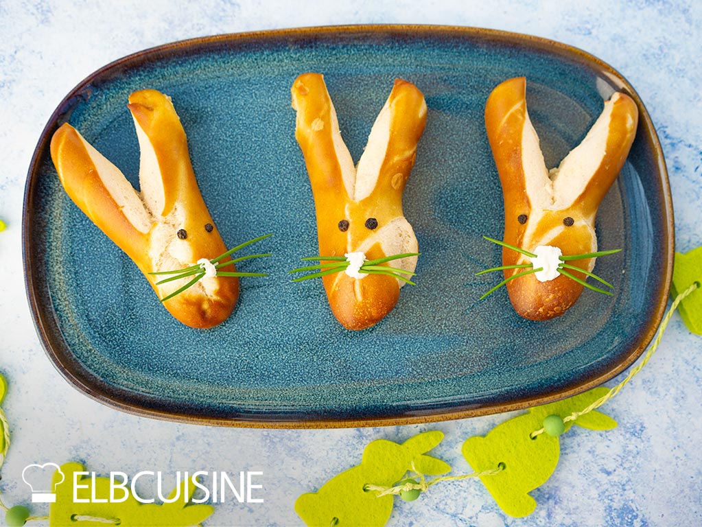 Eine Platte mit goldbraun gebackenen Laugenbrötchen in niedlicher Hasenform mit langen Ohren und einer Nase aus Frischkäse und Schnurrhaare aus Schnittlauch bereit für den Osterbrunch.