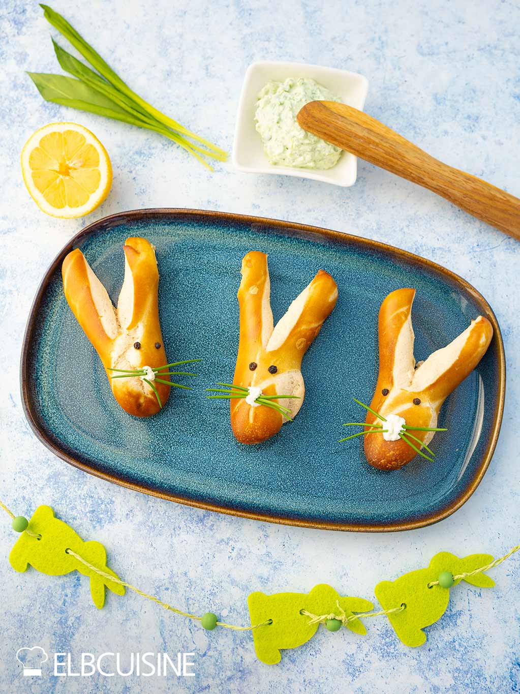 Ein Platte mit goldbraun gebackener Laugenbrötchen in niedlicher Hasenform mit langen Ohren und einer Nase aus Frischkäse und Schnurrhaare aus Schnittlauch mit einem hellgrünen Bärlauch-Dip bereit für den Osterbrunch.